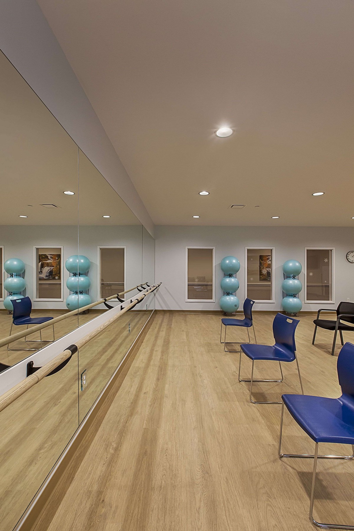 A spacious exercise studio with mirrors, a wooden floor, and blue chairs arranged neatly. Exercise balls are stacked by the wall.
