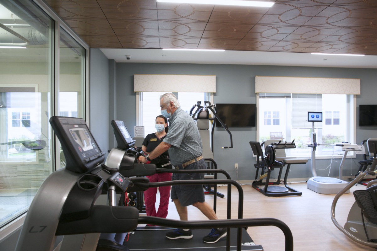 A healthcare worker assists an elderly person on a treadmill in a gym setting.