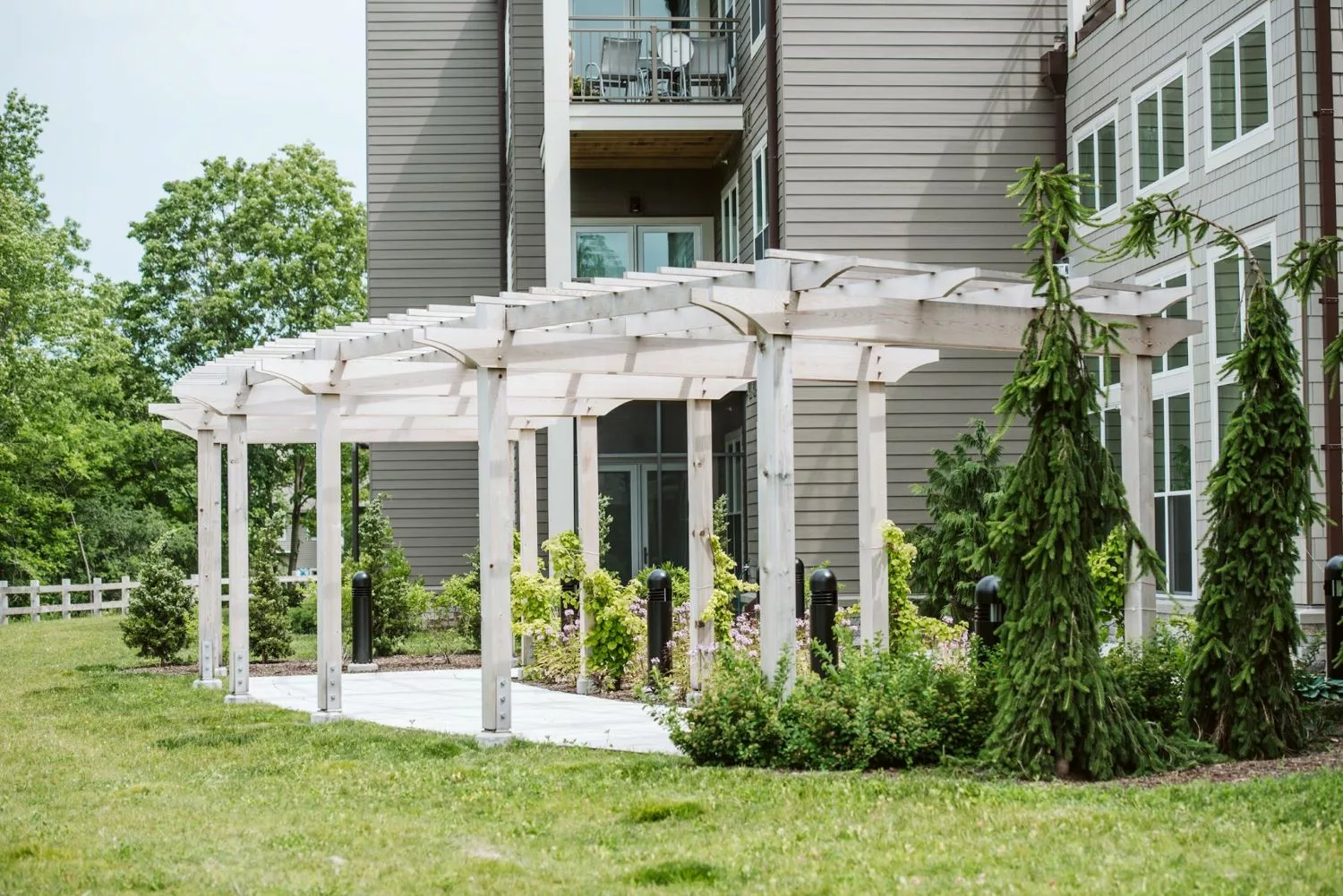 A white pergola in front of a modern apartment building, surrounded by lush greenery.