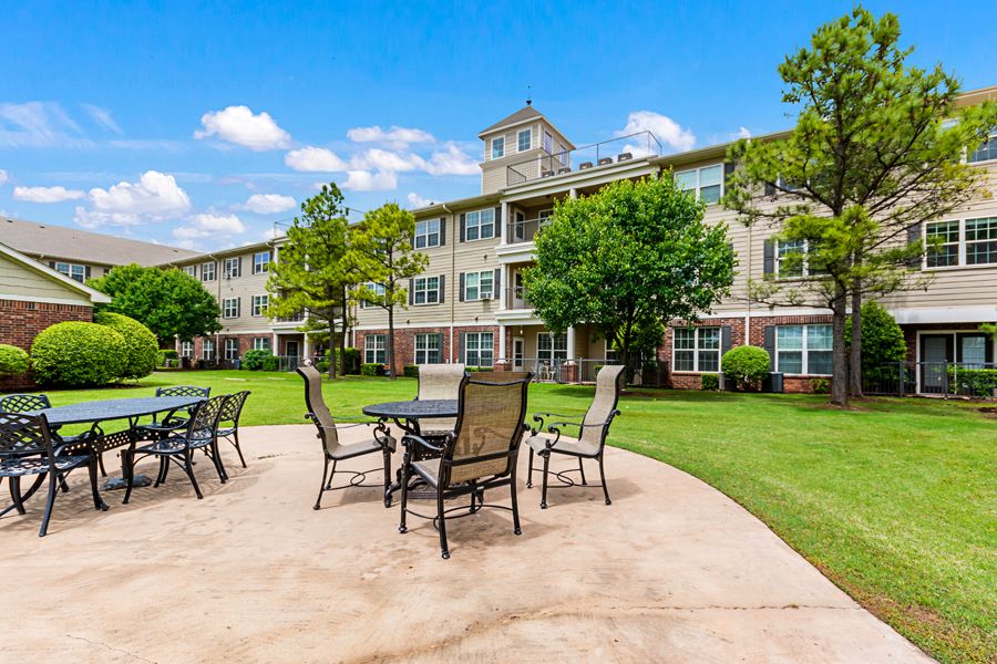 An outdoor patio with tables and chairs, surrounded by a residential building and greenery.