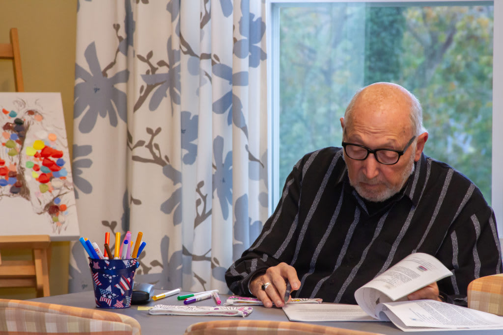 An elderly man wearing glasses is sitting at a table, reading a document with pens and papers around him.
