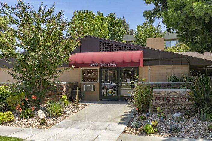 Entrance to a building with a red awning and landscaped surroundings.