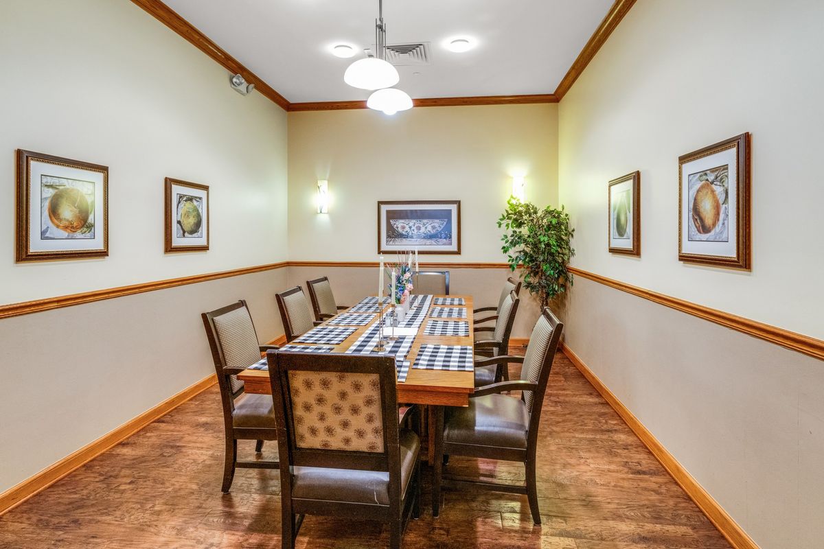 A well-lit dining room with a long table surrounded by chairs, featuring wall artwork and a decorative plant.