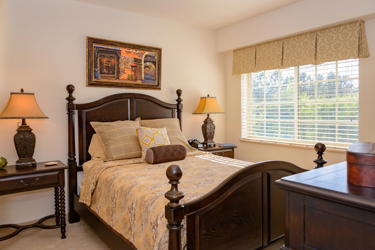 A cozy bedroom featuring a wooden bed, decorative pillows, and a bedside lamp near a window.