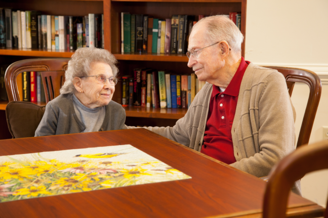 An elderly couple enjoys solving a puzzle together in a cozy room with a bookshelf in the background.