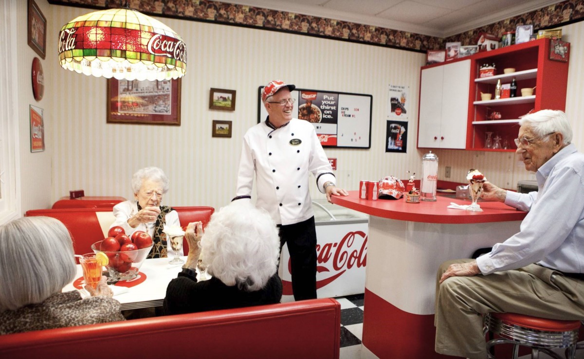 A retro-style diner with a chef speaking to patrons, surrounded by Coca-Cola themed decor.