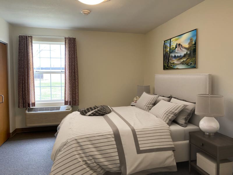 A cozy bedroom with a neatly made bed, featuring neutral-colored bedding and a painting on the wall.