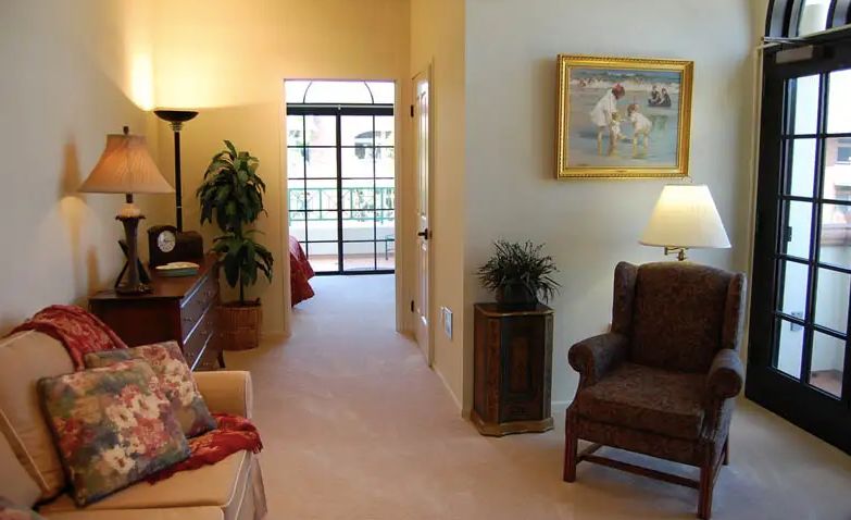 A cozy living room with comfortable seating, artwork on the wall, and indoor plants, leading to a bedroom.