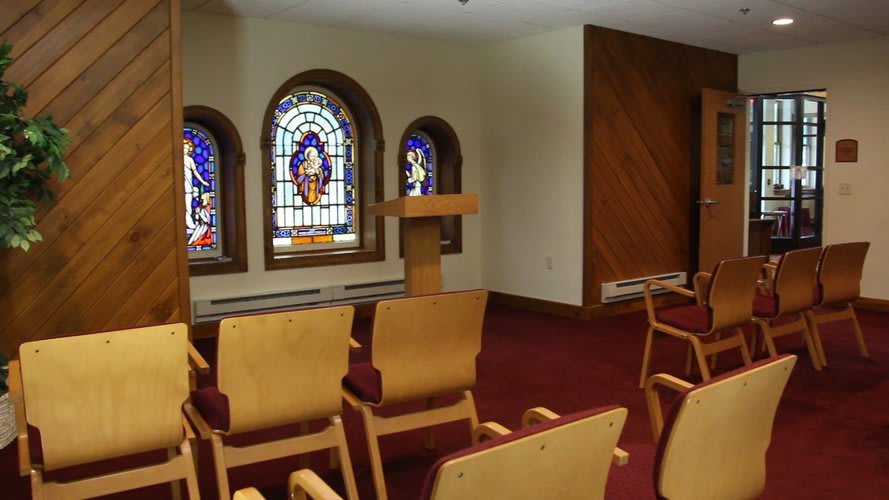 A small chapel interior with stained glass windows, a pulpit, and several chairs arranged in rows.