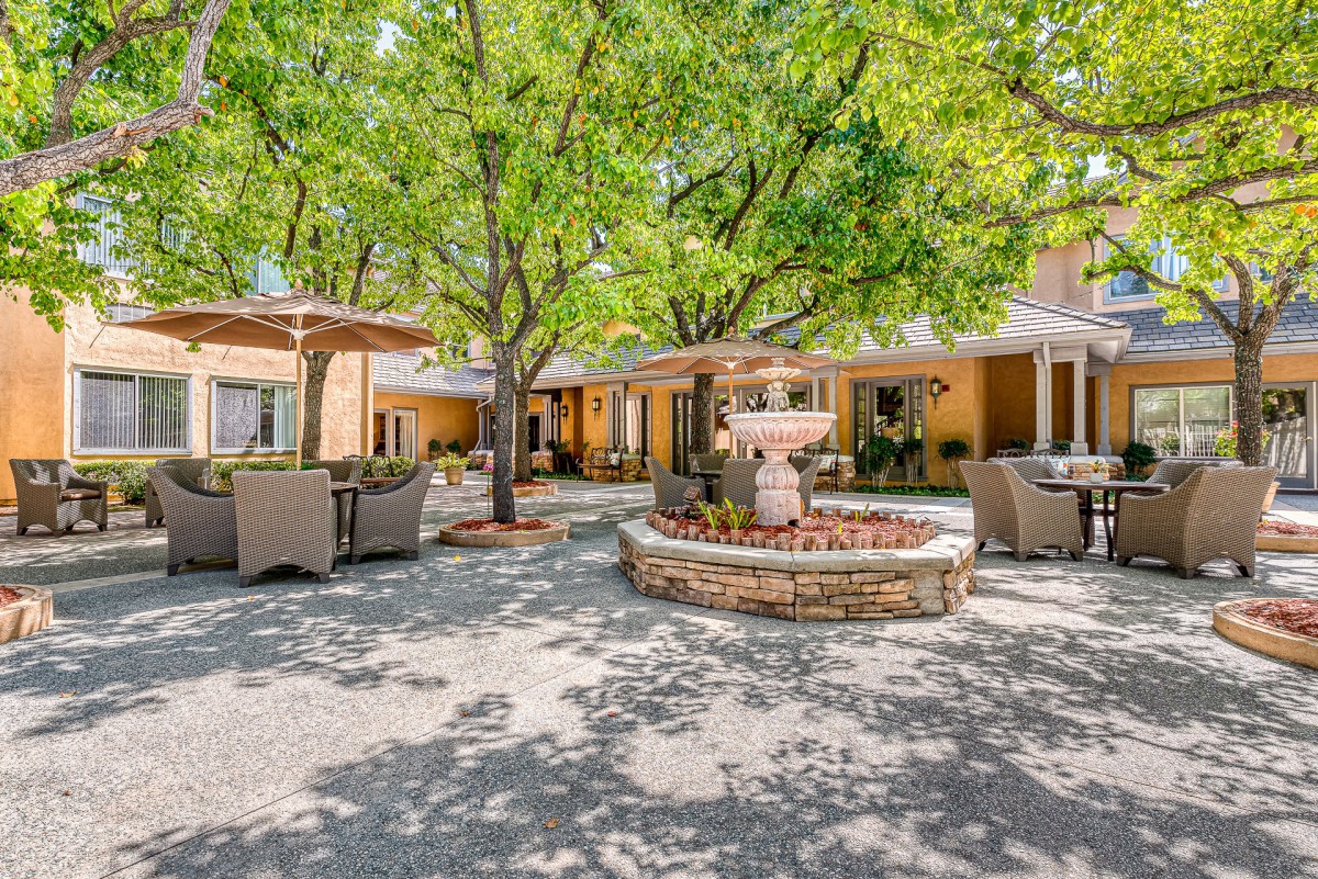 A sunny courtyard with a central fountain, surrounded by outdoor seating and lush trees.