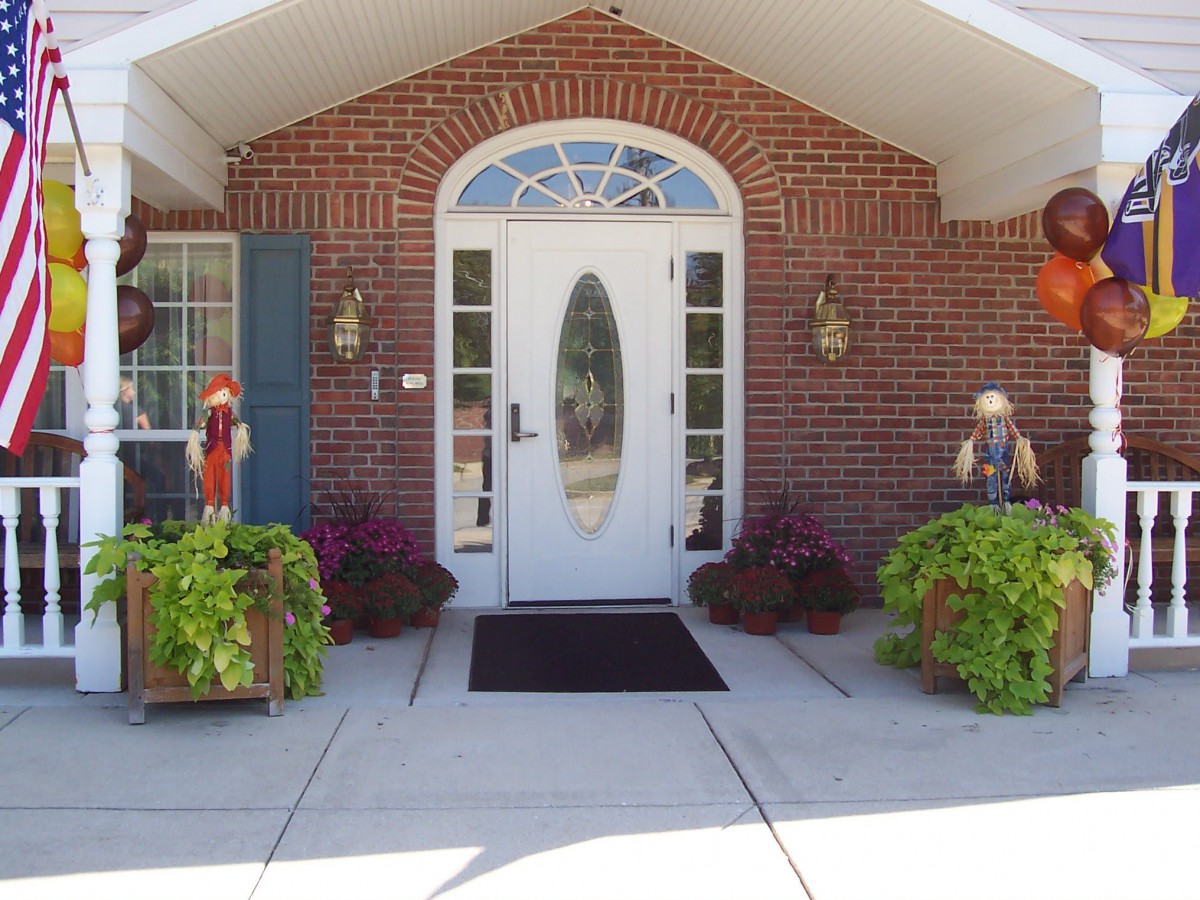 A welcoming entrance with a white, oval-glass front door set in a brick building, flanked by potted plants and outdoor decorations.