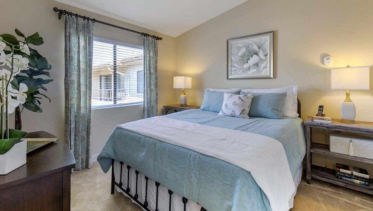 A cozy bedroom with a neatly made bed, light blue accents, and a window allowing natural light.