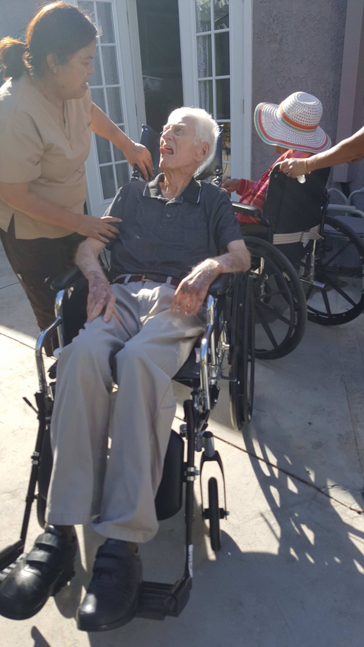 An elderly person in a wheelchair receiving assistance outdoors.