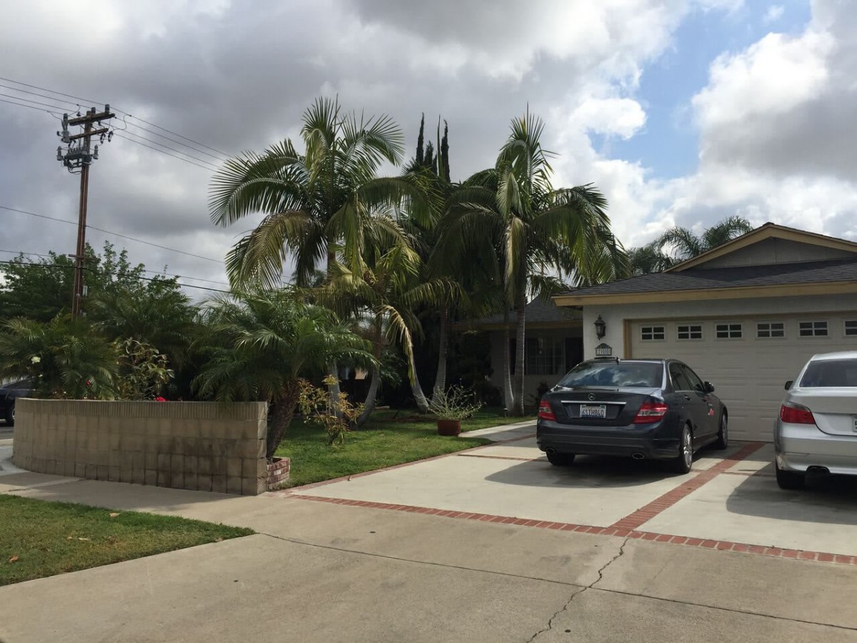 A suburban house with a car parked in the driveway, surrounded by palm trees under a cloudy sky.