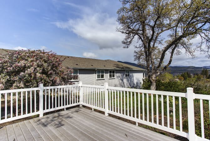 A spacious wooden deck with white railings overlooks a yard with trees and a house in the background under a partly cloudy sky.