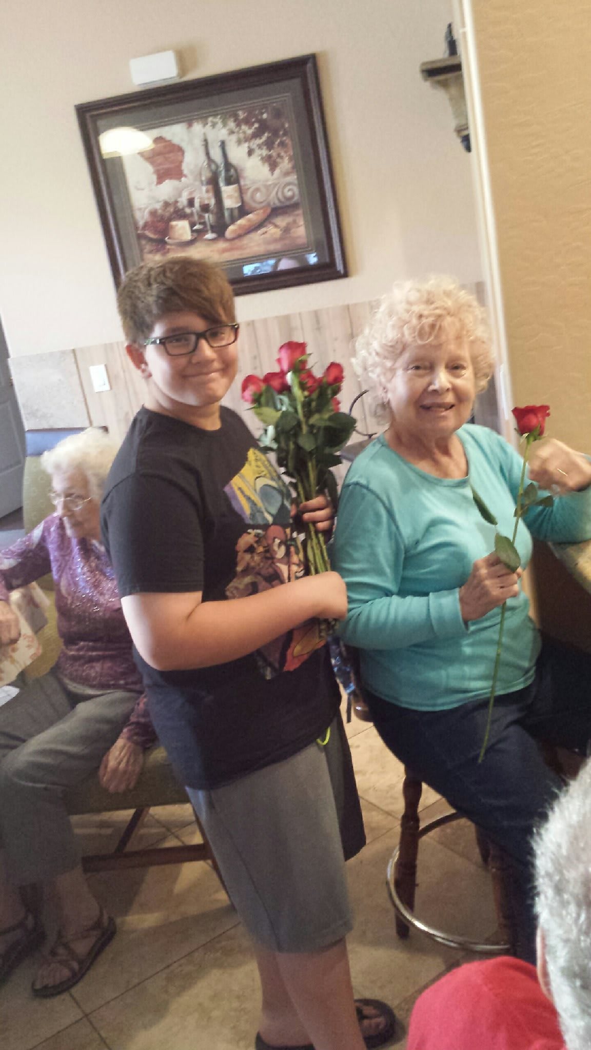 A young boy holds a bouquet of roses while standing beside an older woman, both smiling in a cozy indoor setting.