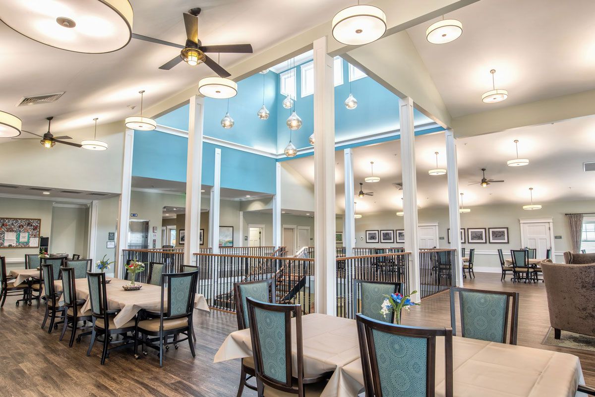 A spacious and bright dining area with modern decor, featuring tables and chairs arranged neatly under a high ceiling with fans.