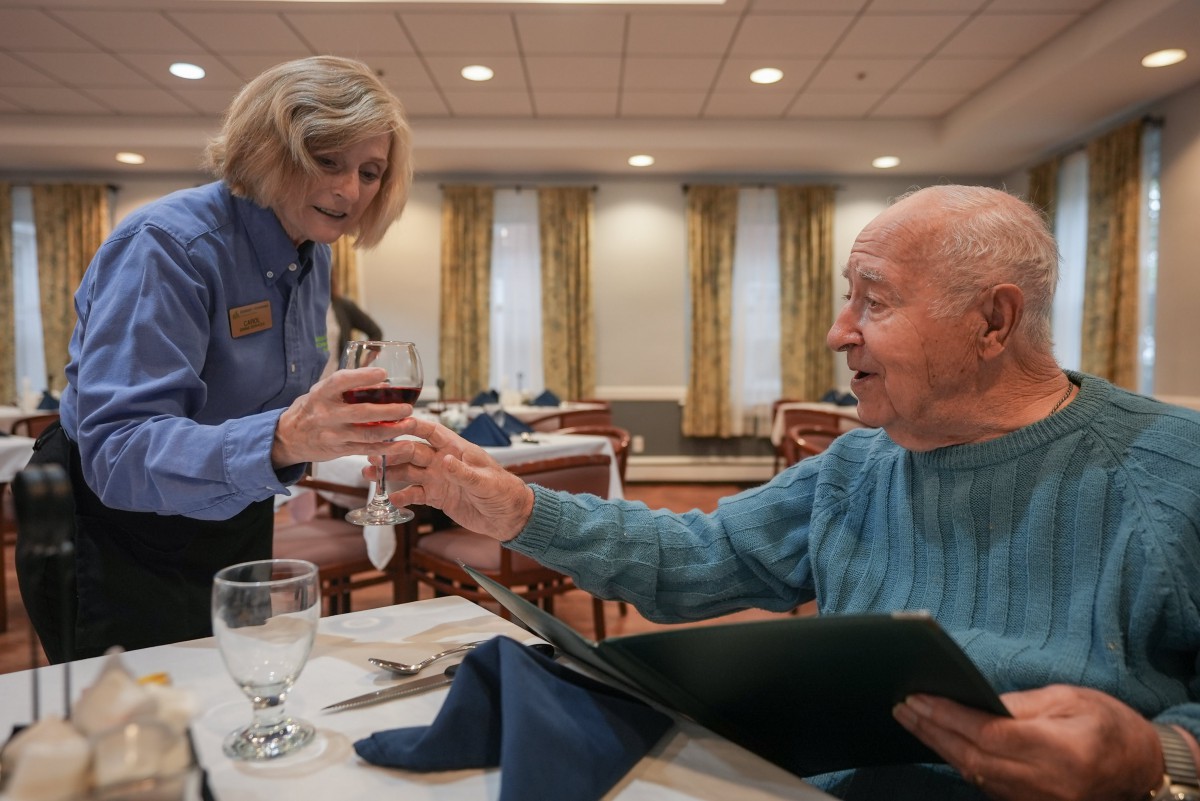 An elderly man enjoying a meal as a waitress serves him wine in a restaurant setting.