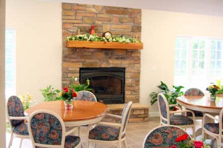 A cozy living room with a stone fireplace and a wooden mantel, adorned with greenery and flowers. Surrounding the round table are chairs with patterned cushions, creating a comfortable and inviting atmosphere.