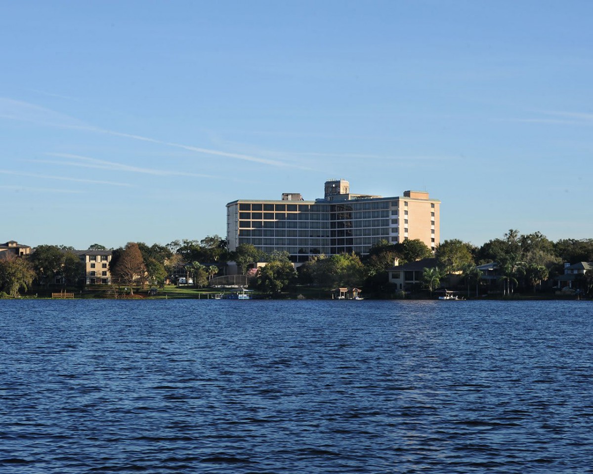 A hotel building overlooking a calm lake surrounded by trees under a clear sky.