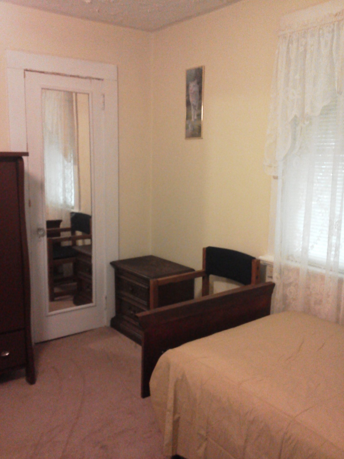 A cozy bedroom featuring a bed, nightstand, mirror, and window with curtains.