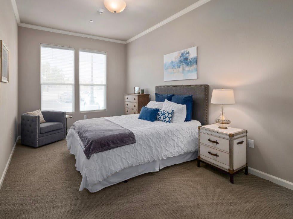 A cozy bedroom featuring a neatly made bed with blue and white bedding, flanked by a nightstand with a lamp. A comfortable chair is placed by the window, and artwork adorns the wall.