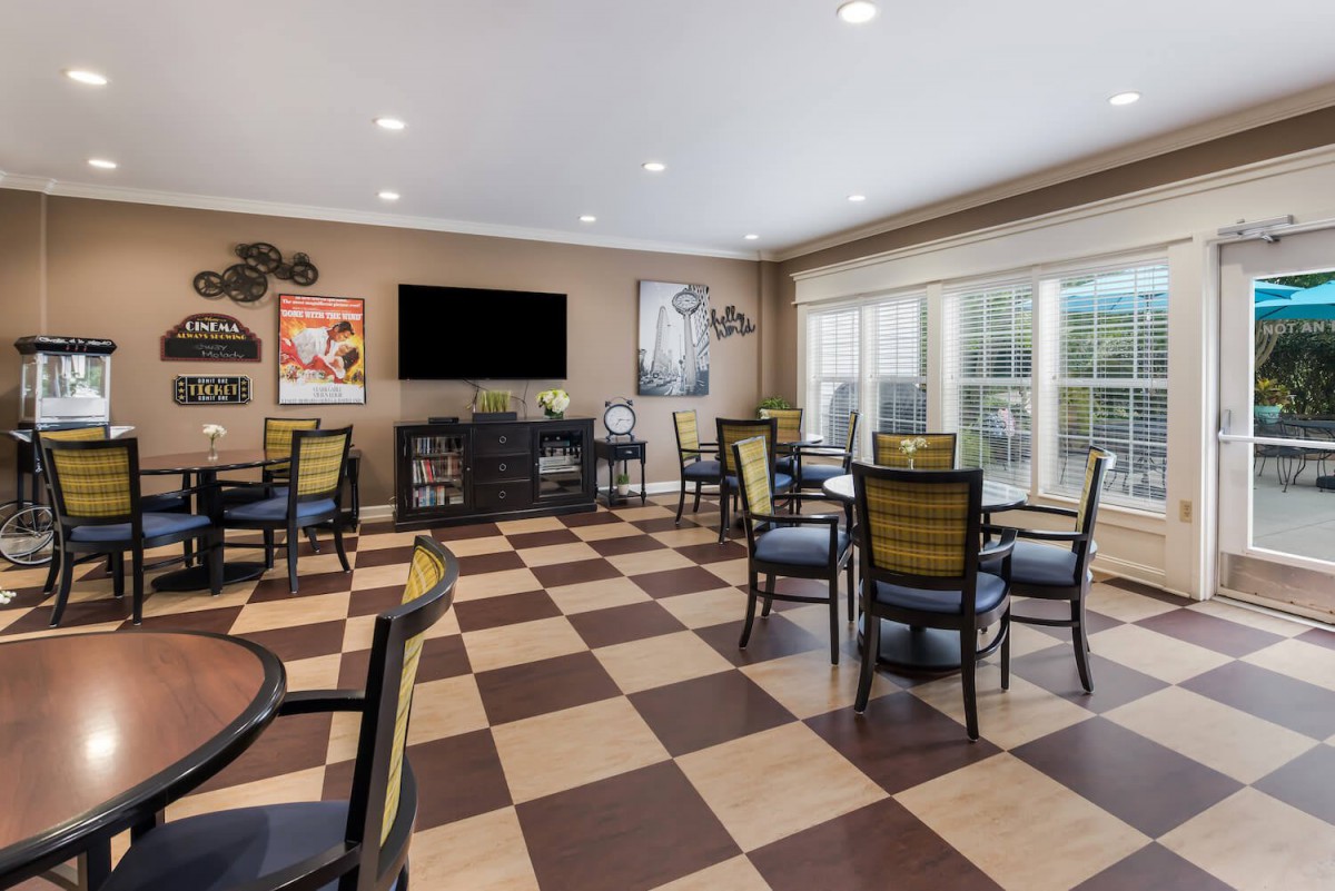 A spacious game room with a checkered floor, featuring several tables and chairs, a large TV, and ample natural light from the windows.