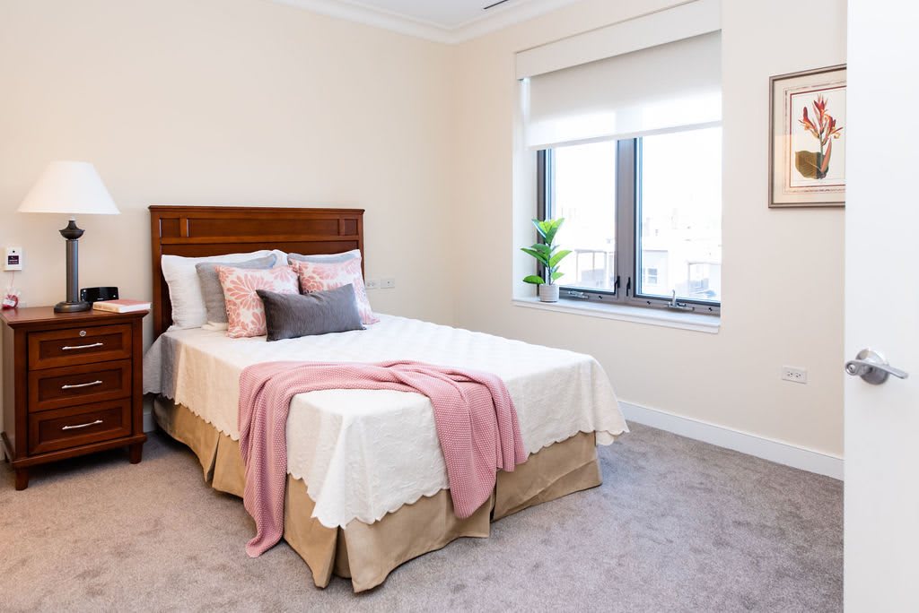 A cozy bedroom featuring a neatly made bed with pink and gray decorative pillows, a soft throw blanket, and natural light streaming through the window.