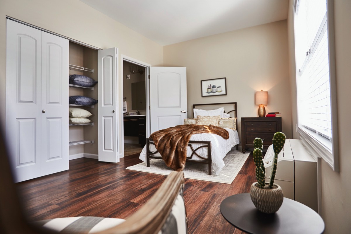 A cozy bedroom with hardwood floors, featuring a neatly made bed, a nightstand with a lamp, and an open closet.
