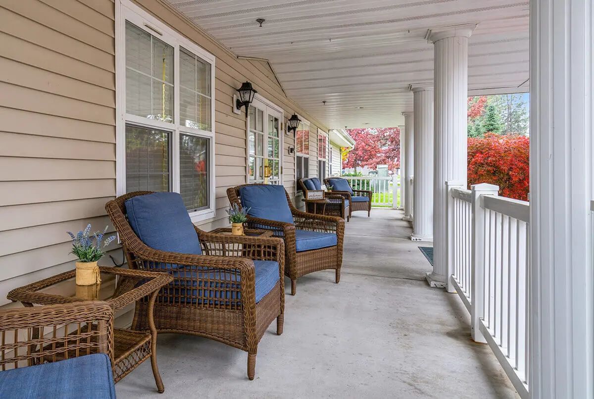 A cozy porch with comfortable wicker chairs and cushions, set against a backdrop of columns and windows.