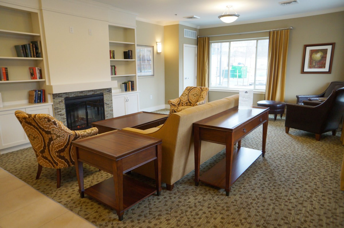 A cozy living room with a sofa, chairs, and a fireplace, featuring a bookshelf and large window.