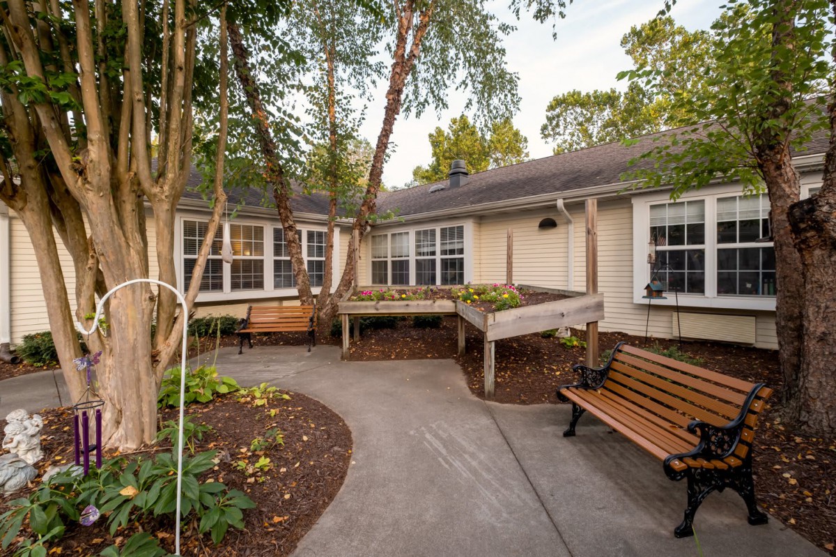 A peaceful garden with a bench and walkway surrounded by trees and landscaping in front of a house.