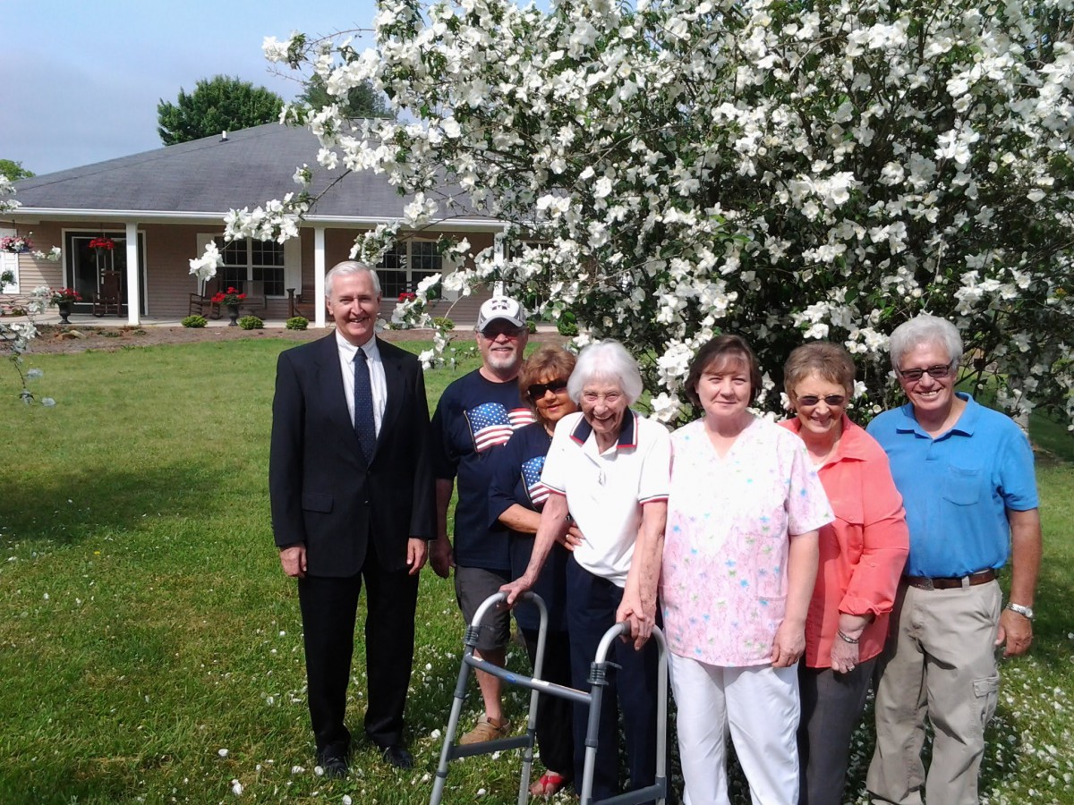 A group of people, including elderly individuals, pose together outdoors near a blooming tree.
