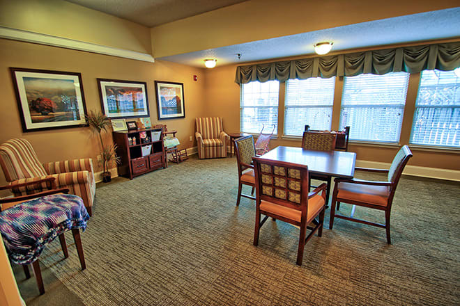 A cozy living room with a table and chairs, carpeted floor, artwork on the walls, and large windows.