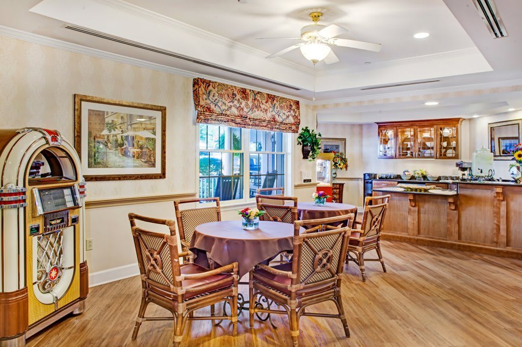 A cozy dining area with a round table and wooden chairs, featuring a nearby kitchen and a window with decorative blinds.