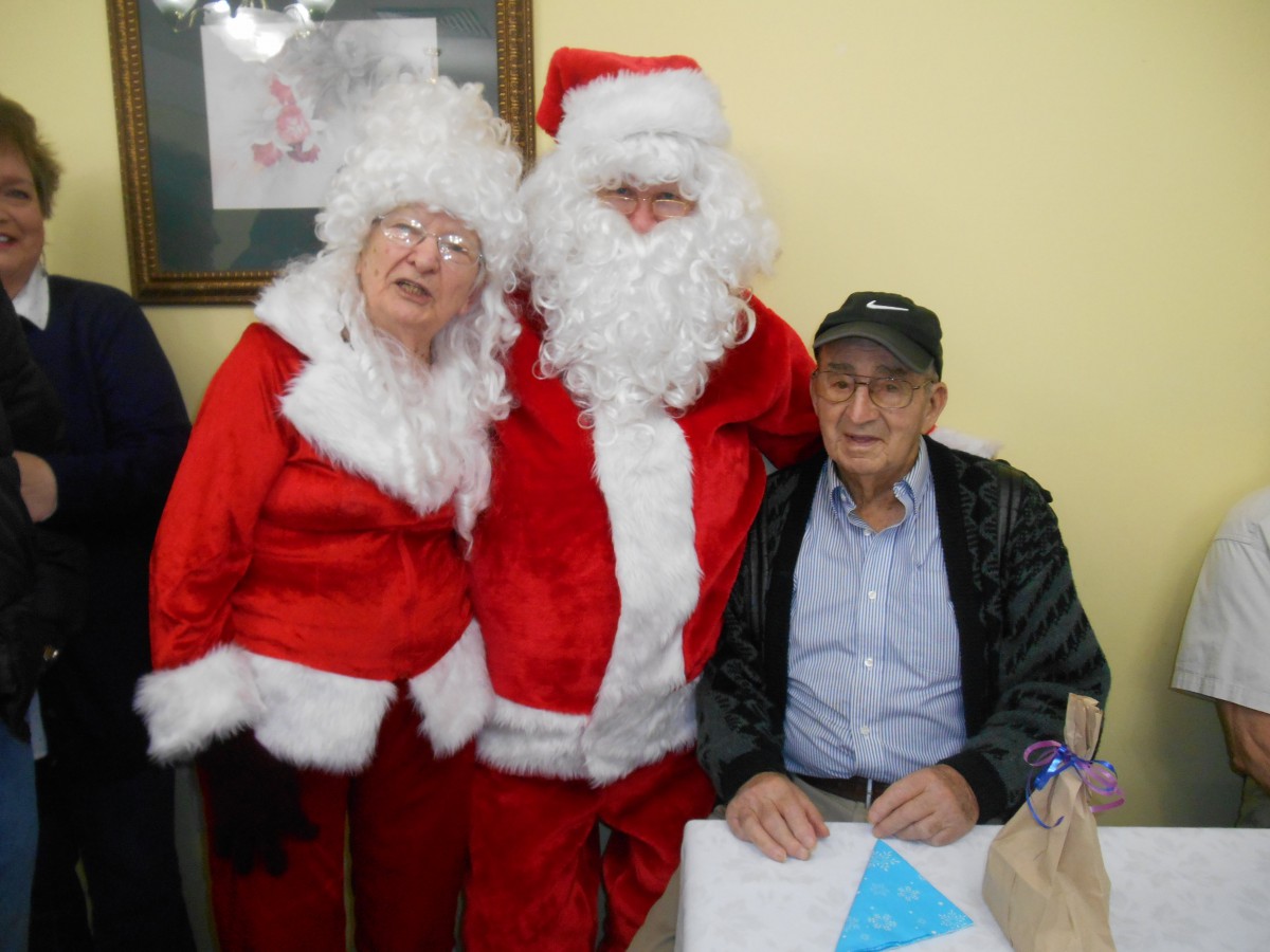 A festive gathering with Santa and Mrs. Claus posing next to an elderly man seated at a table.