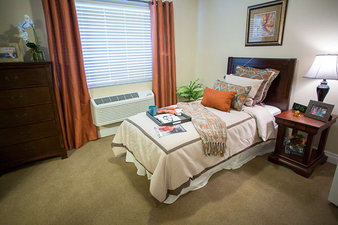 A cozy bedroom with a single bed featuring orange accents, a window with blinds, and an indoor plant.