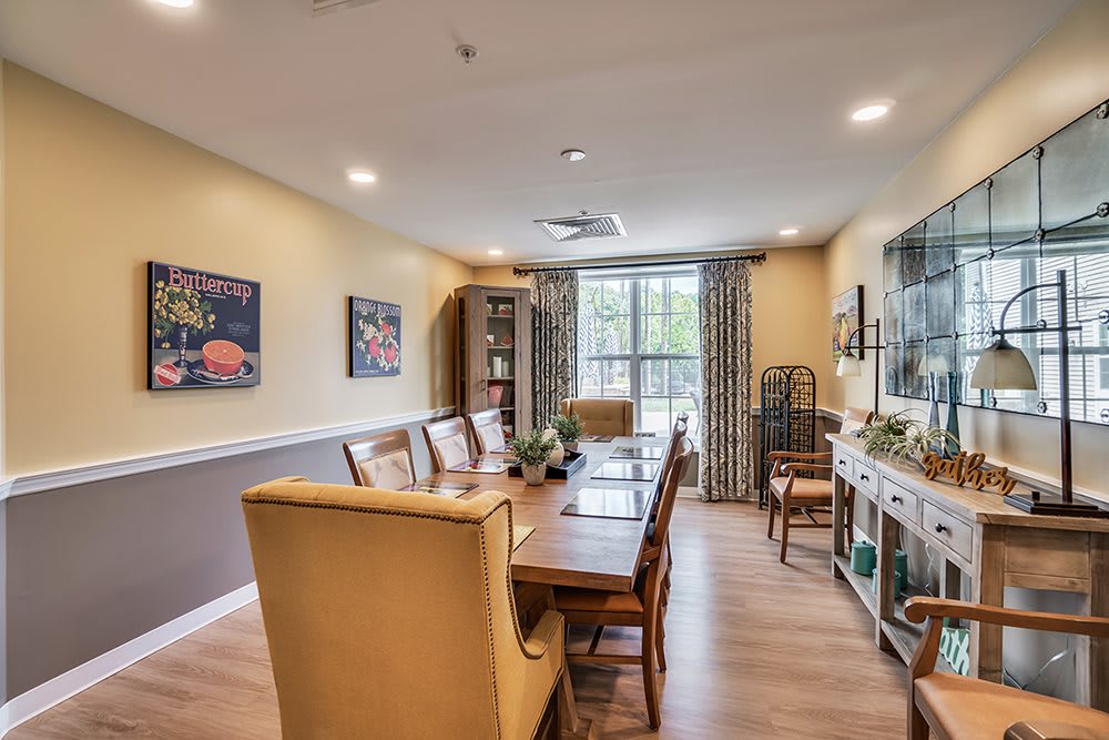 A cozy dining room featuring a long wooden table with chairs, decorated with artwork on the walls and large windows adorned with patterned curtains.