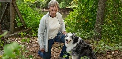 A woman kneeling in a forested area next to a dog, both appearing happy and relaxed.