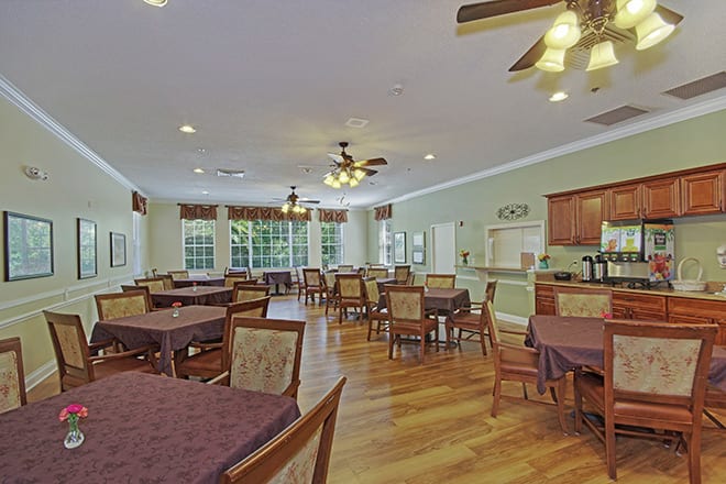 A spacious dining room with multiple tables and chairs, featuring wooden flooring and ceiling fans.