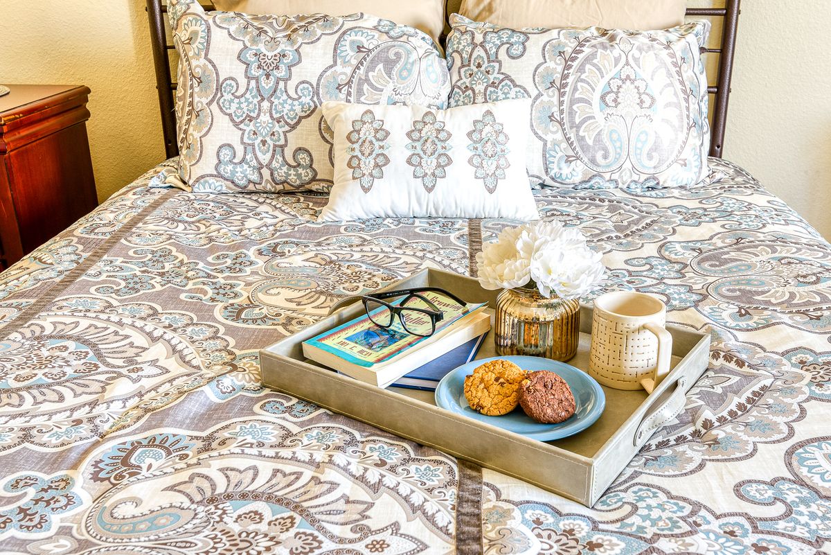 A cozy morning setup with a tray on the bed, featuring a book, glasses, cookies, and a cup of coffee.