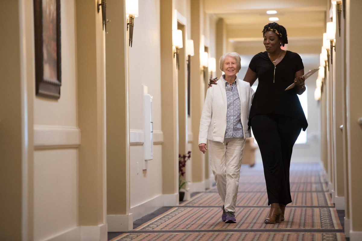 An older adult and a younger adult walking together down a hallway, sharing a moment of companionship.