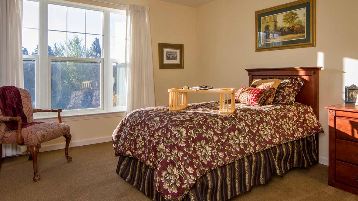 A cozy bedroom featuring a floral-patterned bedspread, with a breakfast tray placed on the bed, and sunlight streaming through the window.