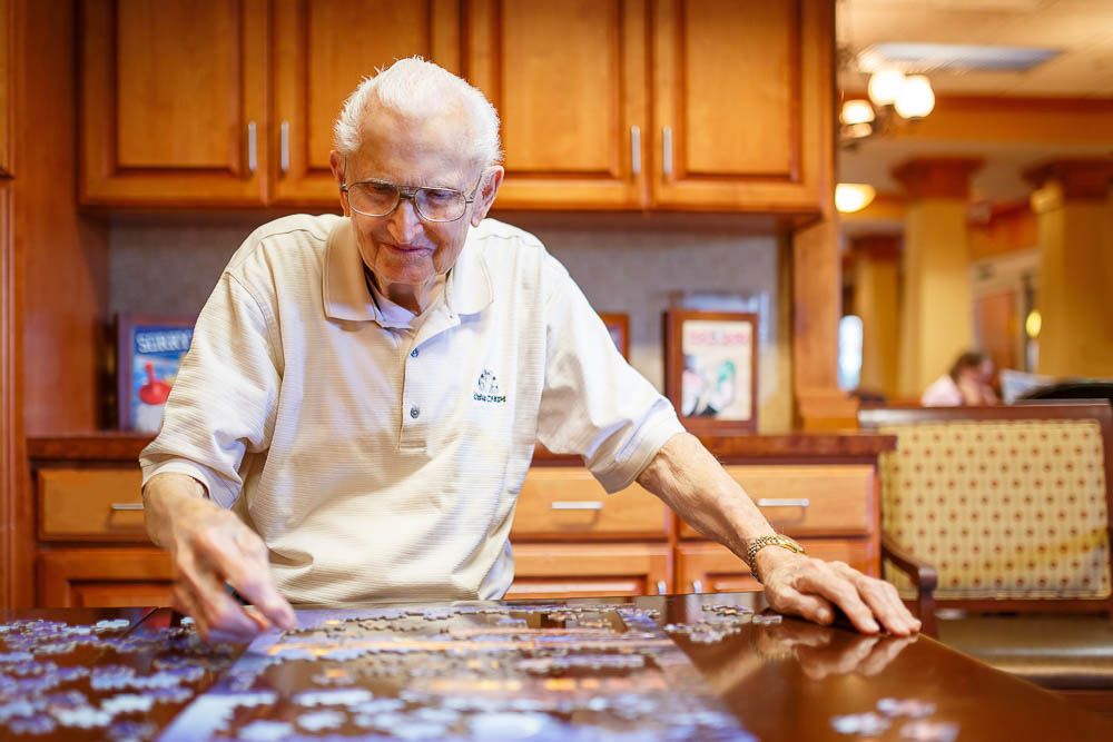 An elderly man happily working on a puzzle at a table in a cozy setting.