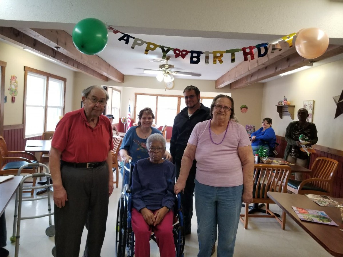 A group of people celebrating a birthday in a community center, with one person seated in a wheelchair and balloons in the background.