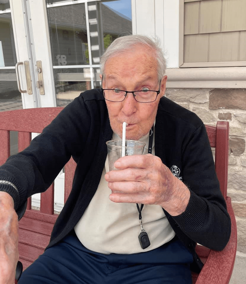 An elderly man wearing glasses is sitting on a bench and drinking from a glass using a straw.