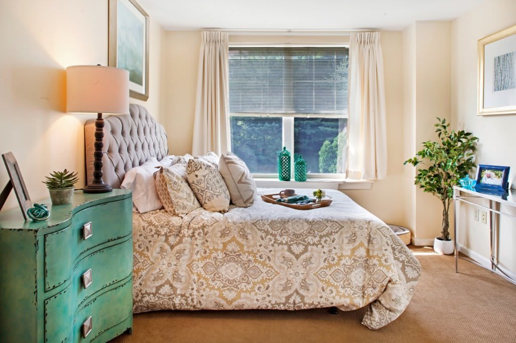 A cozy bedroom with a patterned bedspread, a tufted headboard, and decorative pillows. Natural light streams in through the window, enhancing the room's inviting atmosphere.