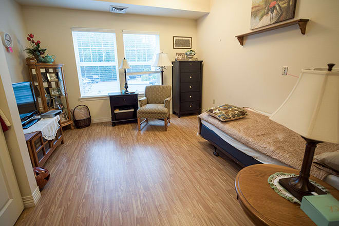 A cozy room with wooden flooring featuring a bed, chairs, a cabinet, and a window allowing natural light.