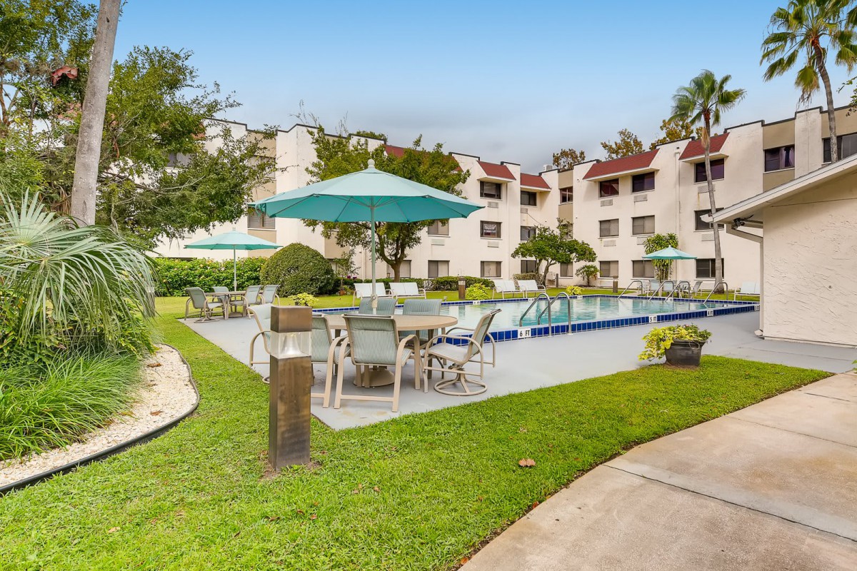 A hotel courtyard with a swimming pool, surrounded by tables and chairs with umbrellas, amidst well-maintained greenery.
