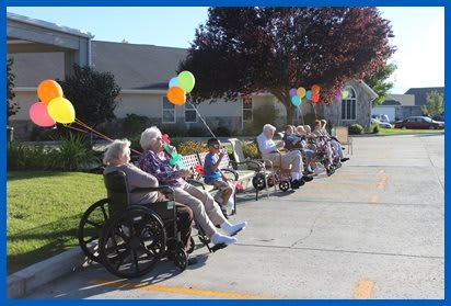 A group of elderly people enjoying a sunny day outside, sitting on benches and in wheelchairs, with colorful balloons around them.